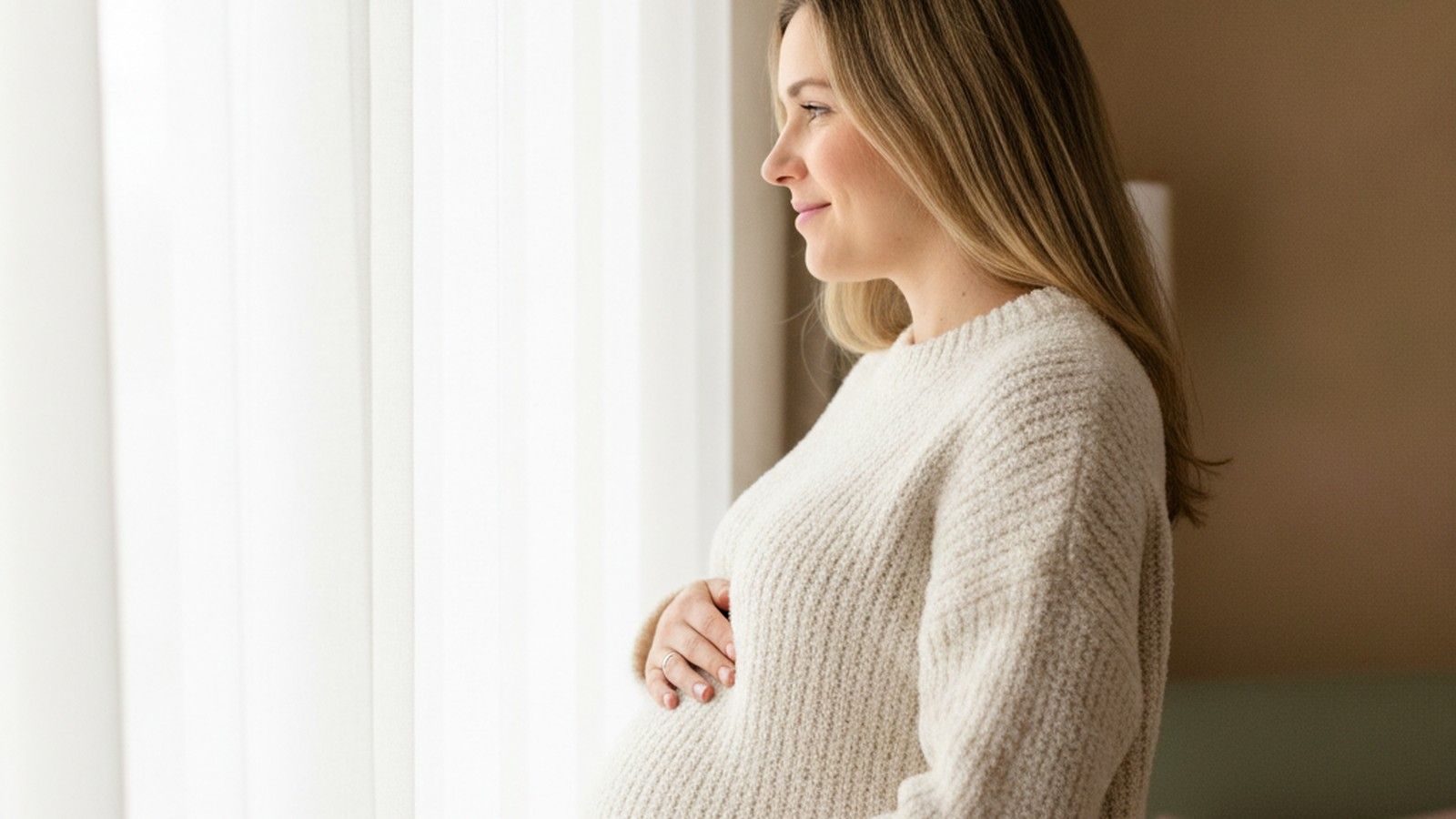 Eine Frau in etwa der zwanzigsten Schwangerschaftswoche steht am Fenster, die Hände auf dem Bauch
